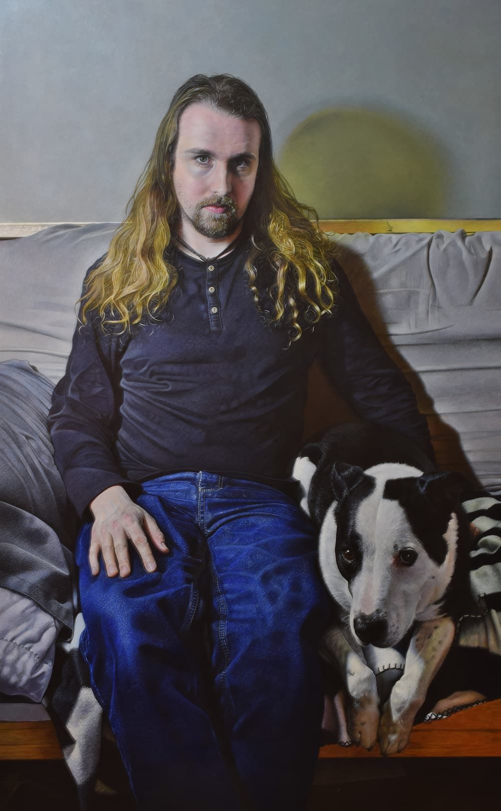 A man with long hair with his hand resting on the black and white do beside him. Both subjects are looking forward.