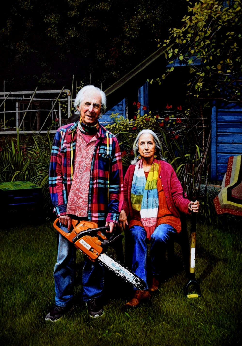 A man holding a chainsaw standing next to a seated woman holding a gardening fork in brightly coloured clothes in a garden setting