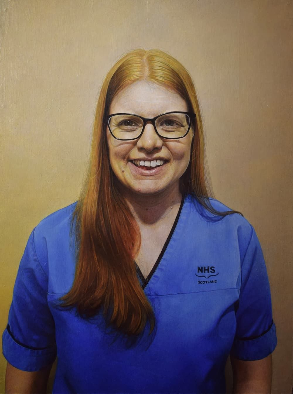 A smiling red-haired woman with glasses in a blue NHS nurse's uniform looking straight ahead.