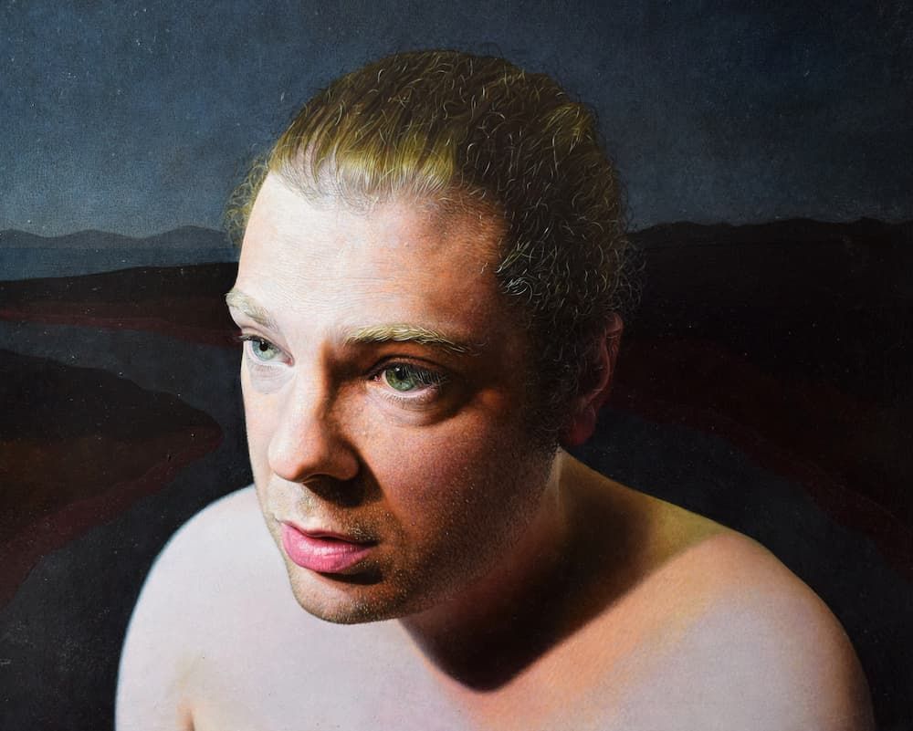 Head and shoulders of a man with his hair tied back. He does not have any clothes on. The background is a dark landscape.